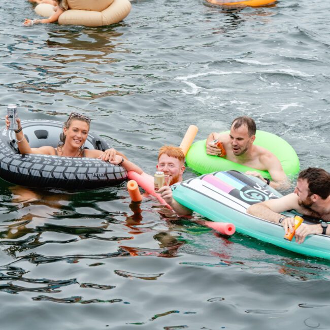 A group of people enjoying a day in the water, lounging on various inflatable floats. They are smiling and appear to be having fun in the lake. Some are holding drinks, and the water is calm, creating a relaxing atmosphere reminiscent of The Yacht Social Club Event Boat Charters.