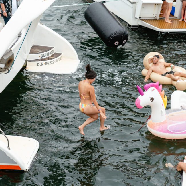 People are enjoying a sunny day on the water. Some are on boats, rented from The Yacht Social Club Sydney Boat Hire, while others are in the water on large inflatables, including a unicorn float. One person is mid-air, having just jumped in. The atmosphere is lively and festive.