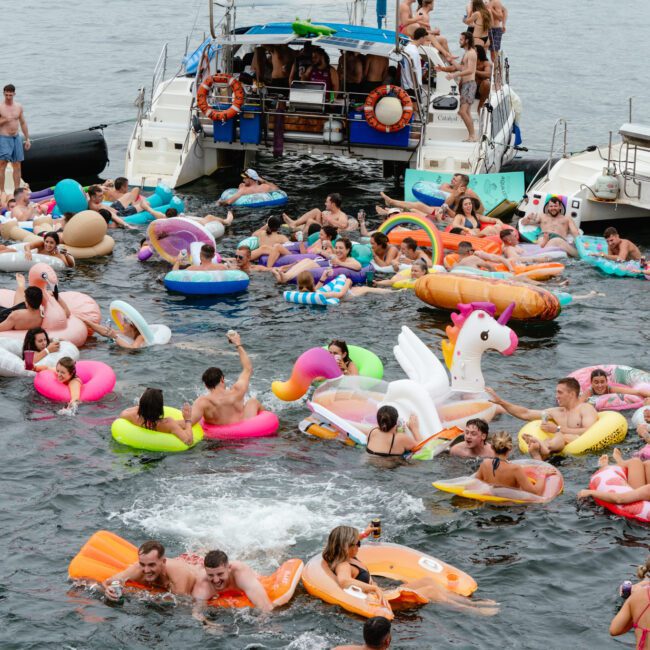 A large crowd of people enjoying a party in the water, surrounded by colorful inflatable floats shaped like animals and other fun designs. Various yachts from the Boat Parties Sydney The Yacht Social Club are docked nearby. The atmosphere is lively with people laughing, talking, and having fun.