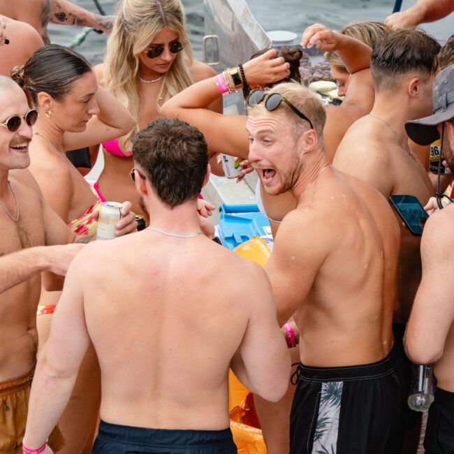 A group of people are gathered closely together, enjoying a lively outdoor event. Many are shirtless, wearing swimsuits or summer attire, and holding drinks. They look cheerful and engaged in animated conversation near a body of water, possibly taking part in The Yacht Social Club's Sydney Harbour Boat Hire.