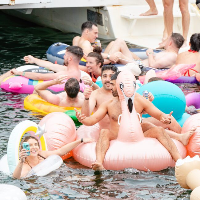 A lively pool party scene where numerous people are enjoying themselves in the water. Some are on colorful inflatable floats, including a large pink flamingo. Everyone appears to be having fun, with one person smiling and holding a drink—much like the vibe at The Yacht Social Club Event Boat Charters.