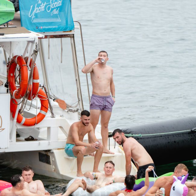 A group of men enjoys a boat party on the water. Several are on an inflatable raft, while others remain on the boat. One man stands on the deck, drinking from a can. The scene exudes a relaxed atmosphere filled with fun and recreation, typical of events hosted by The Yacht Social Club in Sydney Harbour.