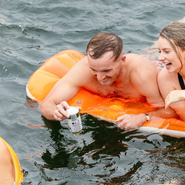 Two adults, a man and a woman, are having fun on an orange inflatable float in the water, holding drinks and smiling. Another person, partially visible, is swimming nearby, adding to the cheerful scene – it’s part of The Yacht Social Club Event Boat Charters experience.