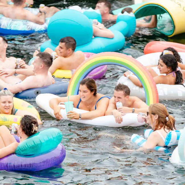 A joyful group of people float in a body of water on colorful inflatable rafts and inner tubes. Some are smiling, laughing, and chatting with each other. Inflatable devices include rainbows, stripes, and various shapes. Sunlight brightens the festive scene organized by Boat Parties Sydney The Yacht Social Club.