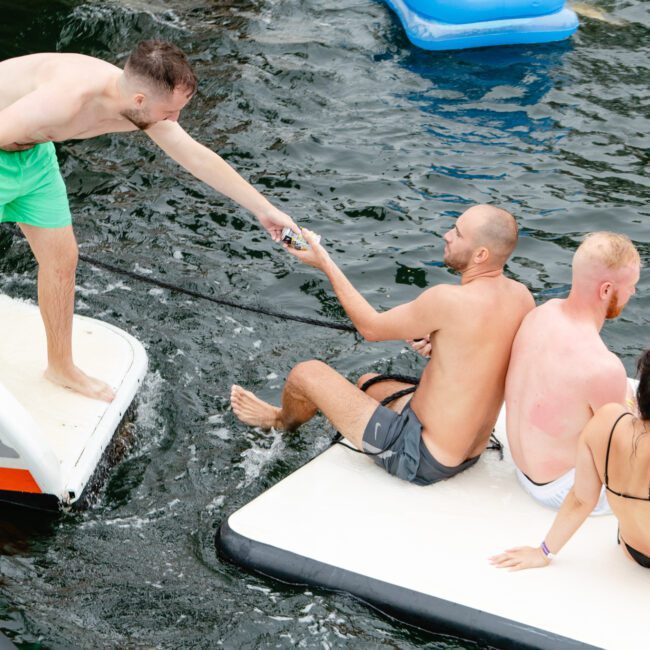 A man in green shorts stands on a boat from The Yacht Social Club, handing a drink to another man sitting on a float in the water. Nearby, three other people relax on inflated water loungers, enjoying the aquatic environment. One person reclines in a blue floating chair.