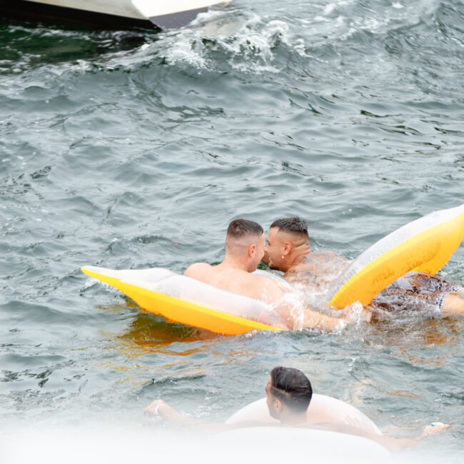 Two men lie on yellow floaties in a body of water, facing each other and appearing to kiss or lean in closely. Another person is partially visible in the lower part of the image, floating in the water. A boat edge from The Yacht Social Club Sydney Boat Hire is visible in the background.