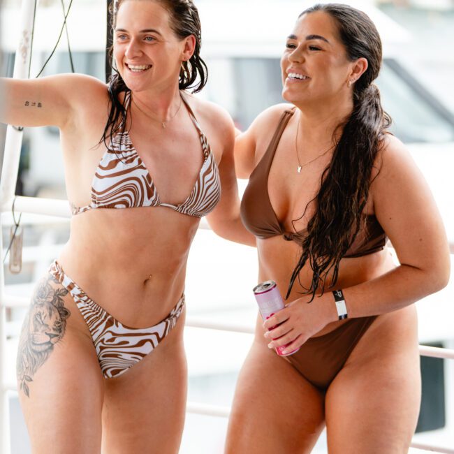 Two women in bikinis smile and pose together on a luxury yacht rental in Sydney. One wears a patterned bikini and has tattoos; the other is in a solid-colored bikini, holding a beverage can. The background shows a blurred marina with boats and water, capturing the spirit of The Yacht Social Club's boat parties.