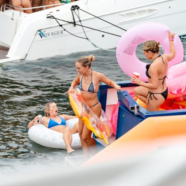 Three women are enjoying a sunny day on the water near a boat named "Voyager." One woman floats on an inflatable ring, another prepares to step onto a waterslide, and the third, holding a cup, stands nearby with colorful inflatables. It’s the perfect day for Luxury Yacht Rentals Sydney.