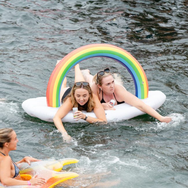 Three people enjoying the water: two women lying on a rainbow-themed inflatable raft, one holding a drink, and another woman relaxing on a star-shaped float nearby. The scene captures a fun and lively moment in a body of water, perfect for those looking into Luxury Yacht Rentals Sydney.
