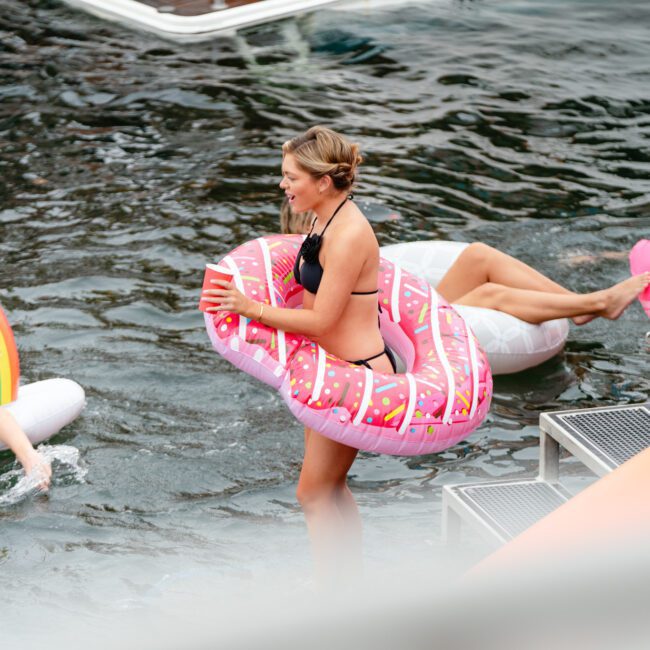 Three people are enjoying time in the water with inflatable tubes. One person in a pink donut float walks up a staircase onto a boat, while another person on the left rests on a rainbow float. A third person in the background floats on a white tube, all part of Sydney Harbour Boat Hire The Yacht Social Club.
