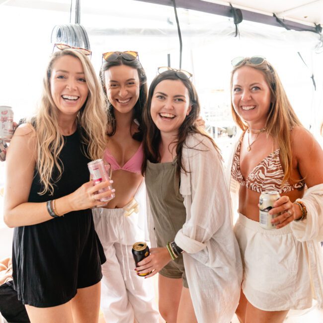 Four women are smiling and enjoying drinks on a boat. Three of them are wearing light, casual summer outfits, while one is in a pink bikini top and white pants. They are under a canopy with sunlight filtering through, suggesting a sunny day out on the water—courtesy of The Yacht Social Club Sydney Boat Hire.
