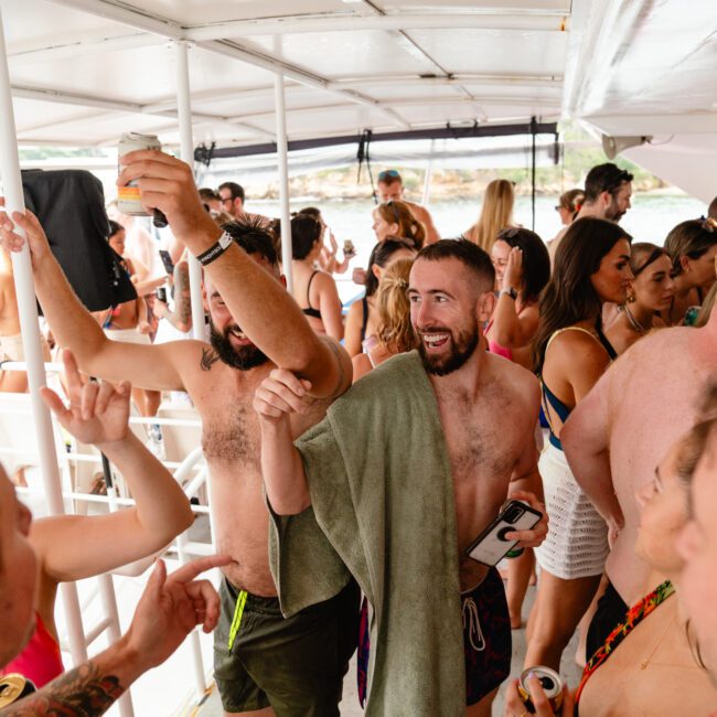 A group of people in swimwear are gathered on a boat, mingling and enjoying drinks. One man with a towel around his shoulders is raising a can and smiling at another man, while others engage in conversation. The atmosphere is lively and social, epitomizing the fun of Boat Parties Sydney with The Yacht Social Club.