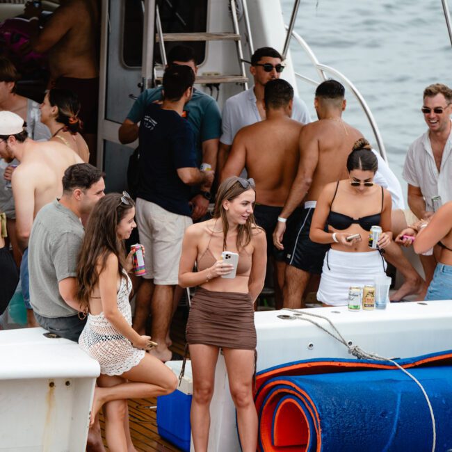 A group of people enjoying a boat party hosted by The Yacht Social Club. Some are standing on the deck, holding drinks and chatting, while others are in swimsuits at the boat's edge. There's a blue foam mat and rolled-up towels on the deck. The background shows beautiful Sydney Harbour waters.