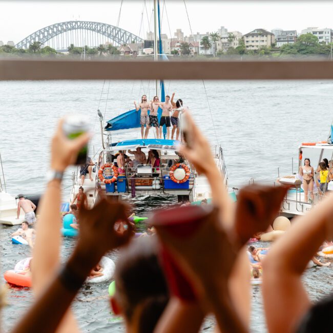Partygoers on boats and inflatables celebrate on a river with the Sydney Harbour Bridge in the background. People on one boat dance and wave, while others in the foreground raise drinks and cheer. The festive scene is lively and joyful, captured perfectly by The Yacht Social Club's Sydney Harbour Boat Hire.
