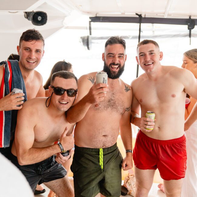 Four men are posing for the camera aboard a boat from Sydney Harbour Boat Hire The Yacht Social Club. Three of them are shirtless, wearing swim trunks, and holding canned drinks. They are smiling and appear to be enjoying a sunny day. Another man and woman are in the background.