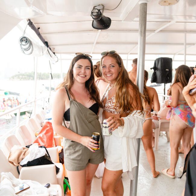 Two women smiling and holding drinks on a luxury yacht. One wears a green romper with a black top, the other in a white outfit with a tan crop top underneath. They are surrounded by others in swimwear, enjoying the outdoor scenery at The Yacht Social Club.