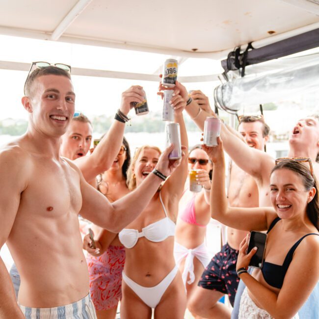 A group of people in swimwear are gathered on a boat, smiling and raising cans and glasses in a celebratory toast. They appear to be enjoying a sunny day with the water visible in the background through the boat's windows, as part of The Yacht Social Club Event Boat Charters.