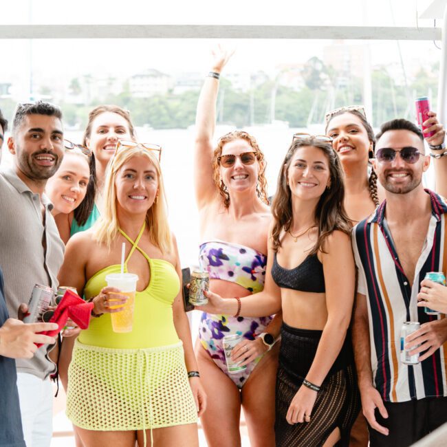 A group of nine people, smiling and posing on a boat. Everyone is dressed in summer attire, including swimsuits, casual tops, and sunglasses. They appear to be enjoying a warm day at The Yacht Social Club Sydney Boat Hire, holding drinks with a scenic backdrop of water and greenery behind them.