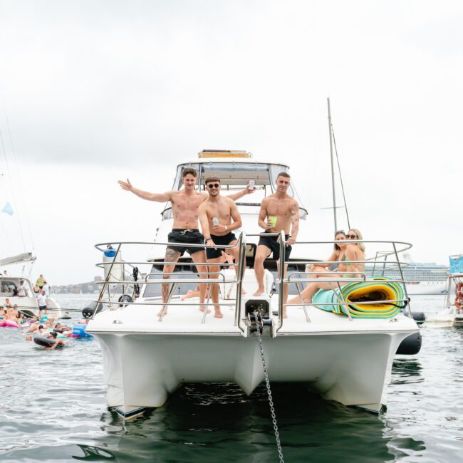 A group of people on a boat enjoying a day on the water. Three men stand shirtless at the bow, smiling and waving. The boat is surrounded by other boats and people swimming or lounging on inflatables. The sky is overcast but the mood appears festive and relaxed, perfect for a Sydney Harbour Boat Hire The Yacht Social Club event.