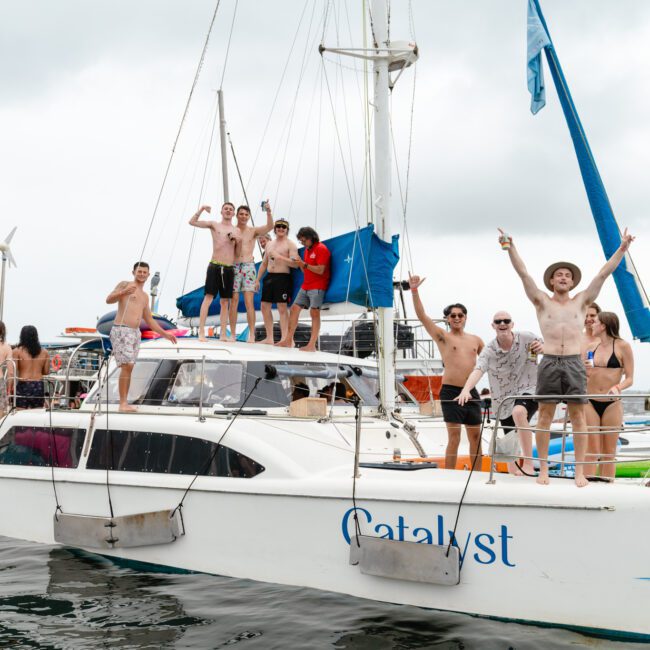 A group of people are on a white sailboat named "Catalyst," celebrating. Some are on the deck, waving their arms, while others are standing on the roof. The background shows more boats and a cloudy sky. The atmosphere is festive and joyful, perfect for Boat Parties Sydney The Yacht Social Club.