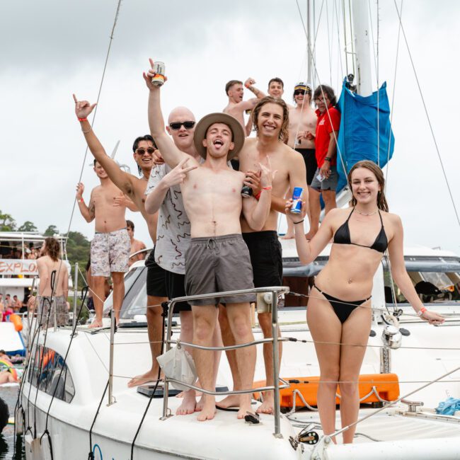A group of people in swimwear stand and celebrate on a sailboat, smiling, holding drinks, and raising their arms in excitement. The Yacht Social Club Event Boat Charters hosts this lively event with other boats visible in the background, creating a festive atmosphere.