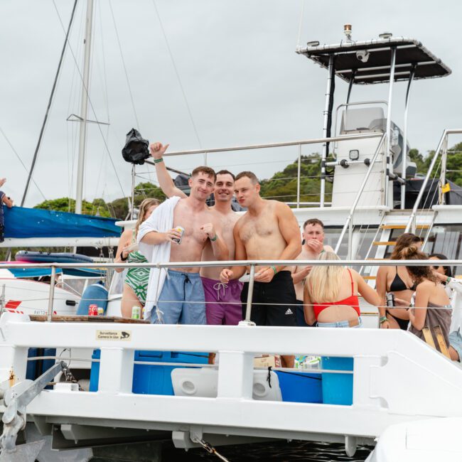 A group of people enjoy a day on a boat. Several men and women, some in swimwear, are standing and socializing on the deck. The weather appears cloudy, and the boat is docked near lush, green foliage—an ideal setting for Sydney Harbour Boat Hire with The Yacht Social Club.