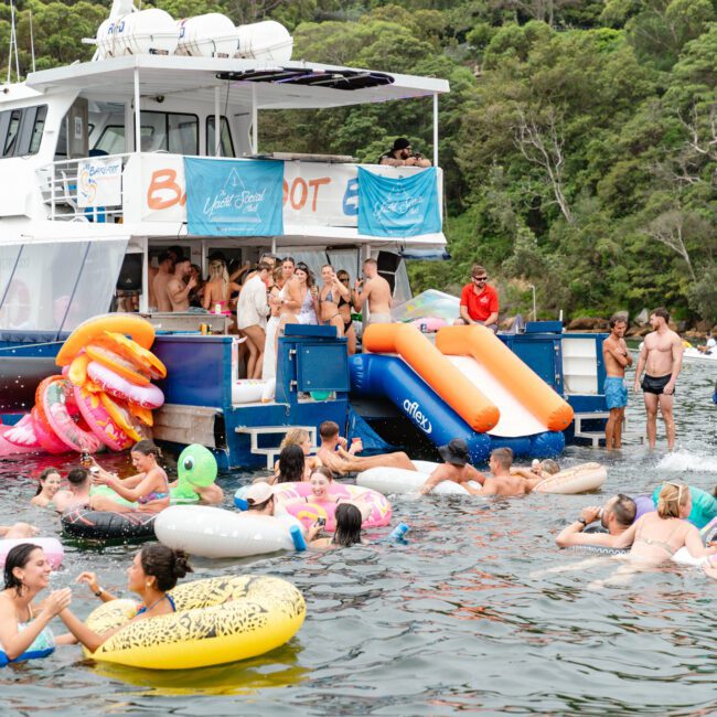 A group of people enjoy a fun day on the water, swimming and floating on various inflatable toys near a blue and white boat. The luxury yacht from Boat Parties Sydney The Yacht Social Club features a slide extending into the water and is anchored near a lush, green shoreline. The atmosphere is vibrant and lively.