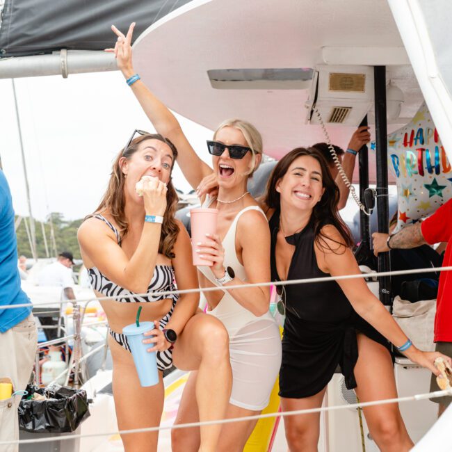 Three women are posing joyfully on a boat. One is standing with her leg raised, another is making a playful gesture with a finger touching her lips, and the third is smiling behind them. Dressed in summer outfits, they appear to be celebrating during a Sydney Harbour Boat Hire The Yacht Social Club event.