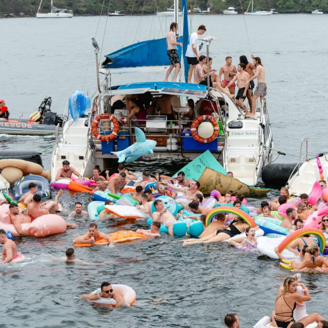 A lively scene of people enjoying a boat party, with many individuals swimming and lounging on colorful inflatables in the water. Several boats are anchored close together with more people onboard, creating a festive and crowded atmosphere. Experience it all with The Yacht Social Club Event Boat Charters.