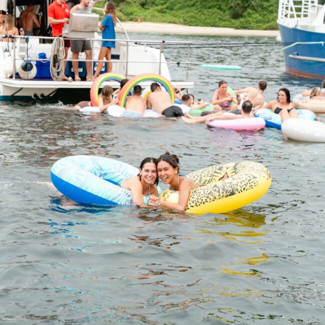 Two women smile while floating on inflatable rafts in the water, surrounded by other people on various inflatables. In the background, boats from The Yacht Social Club Sydney Boat Hire are docked, and people are standing on one of them. It appears to be a lively gathering with a festive atmosphere.
