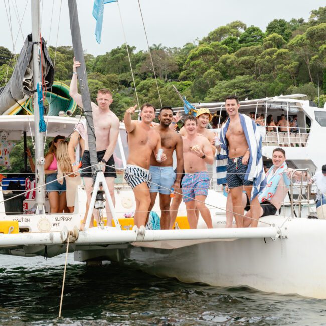 A group of smiling individuals stands on the deck of a yacht, some holding drinks. They are dressed in swimwear and appear to be enjoying a day on the water at The Yacht Social Club Event. Trees and another boat can be seen in the background.