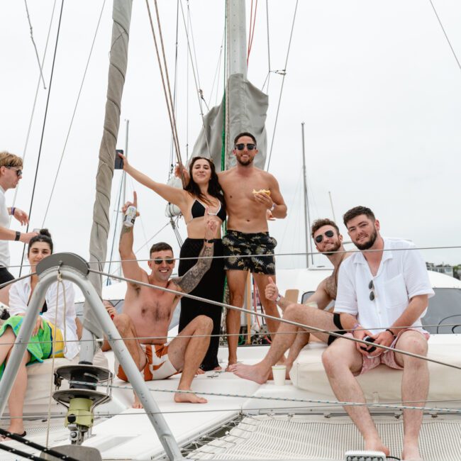 A group of seven people enjoying time together on a sailboat from The Yacht Social Club Sydney Boat Hire. Some are sitting while others are standing, with one person posing with raised arms. They are in swimsuits and casual summer attire, surrounded by sailing equipment and a scenic coastal backdrop.