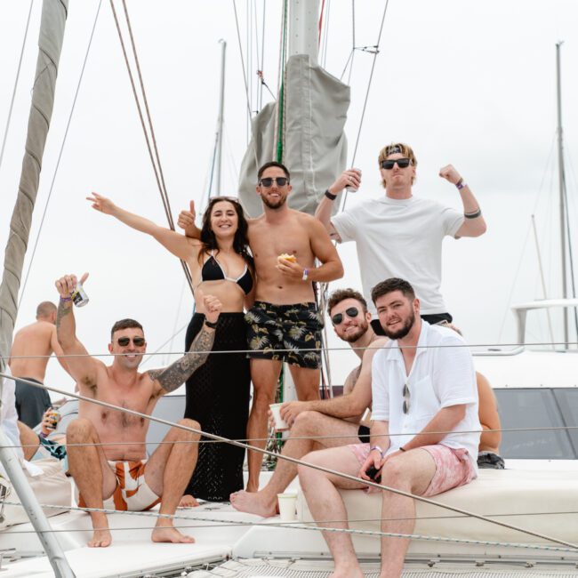 A group of eight people, six men and two women, are posing and enjoying themselves on a sailboat. Some are seated while others are standing, with one woman raising her arms and a man flexing his muscles. The sky is overcast and the mood appears celebratory and fun at The Yacht Social Club Event Boat Charters.