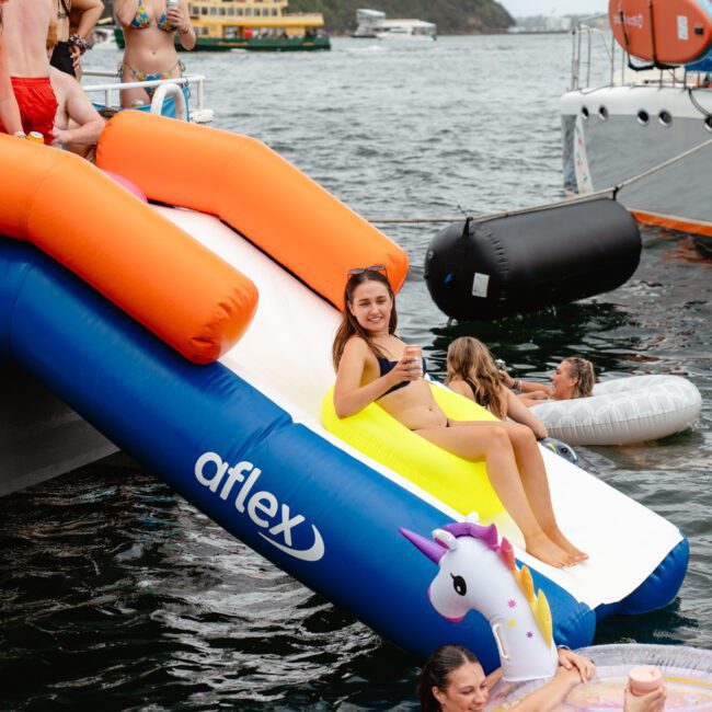 A woman in a bikini slides down an inflatable slide attached to a boat while holding a drink. Other people in swimwear are around her, some in the water and others on the boat. An inflatable unicorn float is in the water nearby, amid a festive atmosphere hosted by The Yacht Social Club Event Boat Charters.