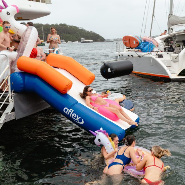 People are enjoying a day on the water with inflatable floats and sliding from a boat into the sea. Several individuals are in the water with unicorn-shaped inflatables, while others are on the boat. The scene is lively and summery, set against a backdrop of boats and shoreline, courtesy of Sydney Harbour Boat Hire.