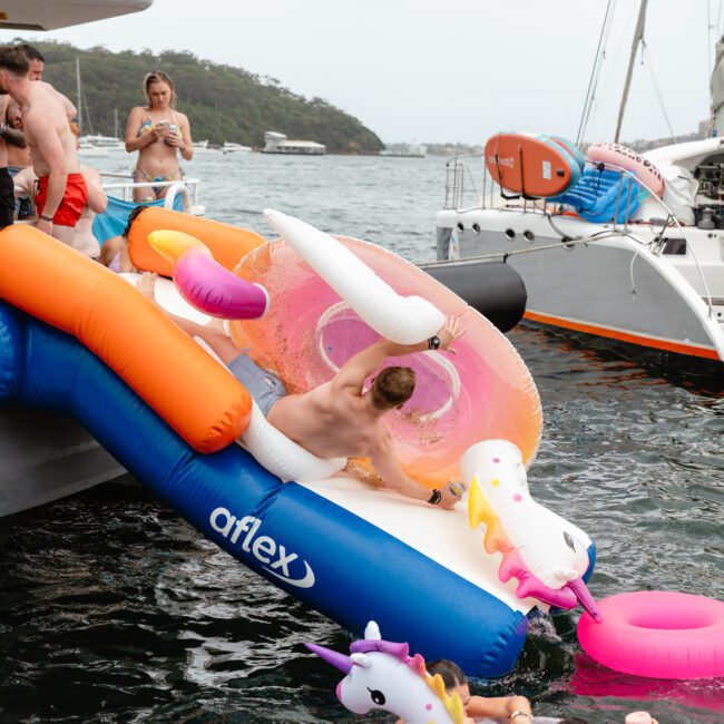 A man slides down a blue inflatable slide into the water, surrounded by colorful pool floats like a pink flamingo and unicorns. Several people on the boat in swimwear watch, while others are in the water. Yachts and a scenic coastline provide a picturesque backdrop at The Yacht Social Club Event Boat Charters.