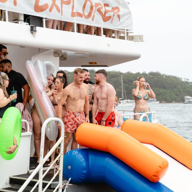 A group of people in swimsuits enjoy a party on a boat, some holding drinks and inflatable pool toys. Two men stand at the top of a slide, while others chat and laugh nearby. The Yacht Social Club Sydney Boat Hire offers such vibrant experiences near scenic waterfronts with trees and boats in the background.