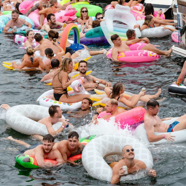 A lively scene of numerous people enjoying a pool party. They are floating on various colorful inflatable rafts and toys in the water, including unicorns, donuts, and flamingos. The atmosphere is festive, resembling Boat Parties Sydney The Yacht Social Club events with individuals smiling, laughing, and socializing.