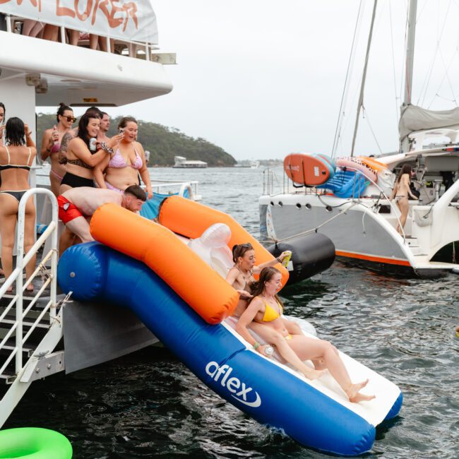 A group of people on a boat in swimwear are sliding off a blue and white inflatable slide into the water. Others watch and wait their turn, with some on nearby boats. The atmosphere is lively, featuring inflatables in the water—typical of Boat Parties Sydney by The Yacht Social Club.