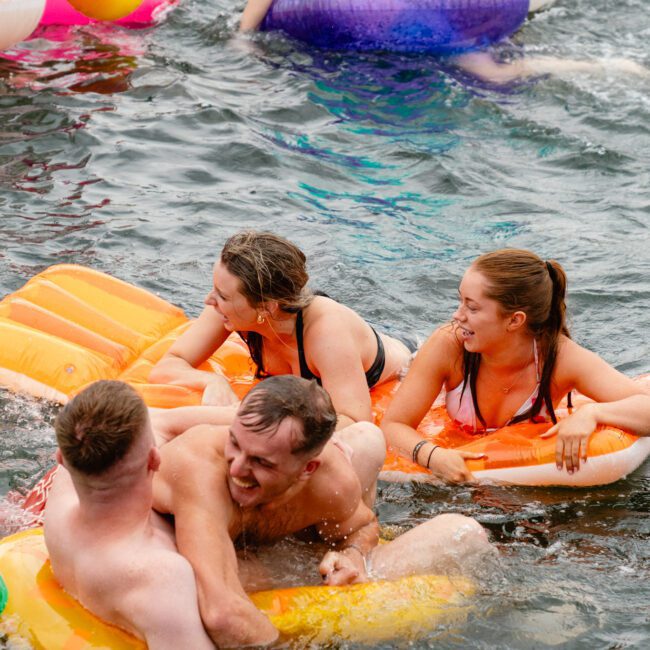 Three people sitting on inflatable pool floats in a body of water are laughing and enjoying a playful moment. One person, half-submerged in the water, interacts with the others. The inflatables are brightly colored, adding to the joyful atmosphere reminiscent of a gathering from Boat Parties Sydney The Yacht Social Club.
