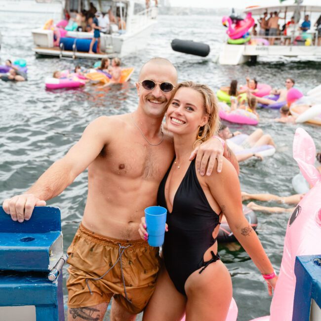 A shirtless man in orange swim trunks and sunglasses stands next to a smiling woman in a black swimsuit holding a blue cup. They are on The Yacht Social Club Sydney Boat Hire boat, surrounded by inflatable pool floats and other people in the water. A large body of water and other boats are visible in the background.