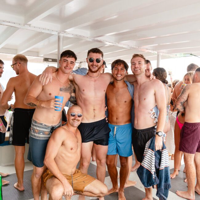 A group of five men pose together on a boat, smiling at the camera. Four are standing and one is crouching in front. They are all shirtless and wearing swim trunks. Other people in swimwear can be seen socializing in the background, enjoying The Yacht Social Club Sydney Boat Hire experience.