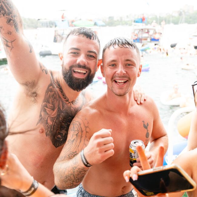 Two men with tattoos and no shirts smile and pose for a photo on a boat from The Yacht Social Club, surrounded by a crowd enjoying a sunny day on the water. They appear cheerful, with one man holding a drink and raising his arm in celebration. Various floatation devices and people are visible in the background.
