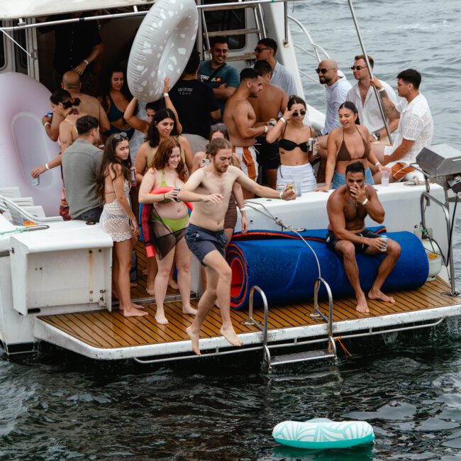 A lively group of people enjoys a boat party on the water with The Yacht Social Club Sydney Boat Hire. Some are on the deck in swimwear, socializing, and others are in the water. Inflatable toys float around, and a man is captured mid-jump off the boat. The atmosphere is festive and fun.