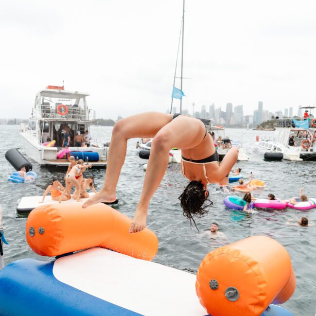 A person in a swimsuit is performing a backflip off an orange and white inflatable structure into the water. In the background, several boats and people swimming on floaties are visible, complemented by a city skyline under a cloudy sky. Luxury Yacht Rentals Sydney creates the perfect backdrop for such adventures.