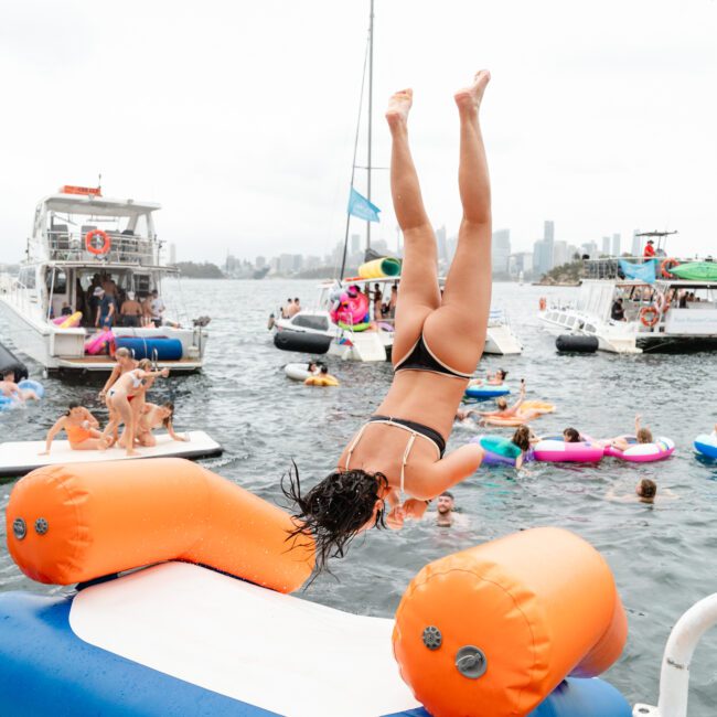 A woman in a black bikini dives off an inflatable water platform into a body of water during The Yacht Social Club Event. Several people on floaties and boats are visible in the background, enjoying a sunny day. The water is calm, and the scene has a lively, recreational atmosphere.