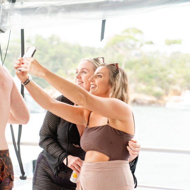 Two women are taking a selfie on a boat. Both are smiling, with the woman on the left holding the phone. They are near water with greenery in the background, enjoying a day out from The Yacht Social Club Sydney Boat Hire. Another person stands to the left, wearing swim trunks.
