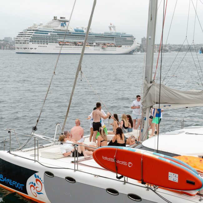 A group of people are gathered on a white sailing catamaran named "Barefoot," with a red surfboard attached to it. In the background, a large cruise ship is seen on the water, and a cityscape is visible under an overcast sky. This scene is perfect for those interested in Boat Parties Sydney The Yacht Social Club.