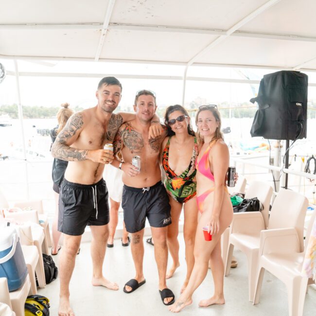 Four adults, two men and two women, are on a boat smiling at the camera. They are dressed in swimwear, holding drinks, and appear to be enjoying a sunny day on the water with The Yacht Social Club Sydney Boat Hire. Other people and water floaties are visible in the background.