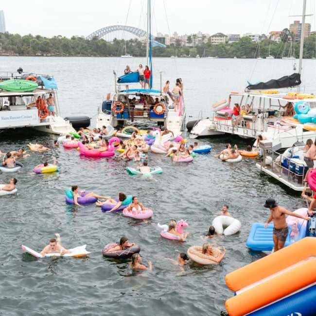 People are enjoying a party on boats and inflatables in a body of water, with Sydney's skyline and the Sydney Harbour Bridge visible in the background. The atmosphere is lively, with colorful floats and boats packed with attendees. It's a festive and summery scene, perfect for Luxury Yacht Rentals Sydney.